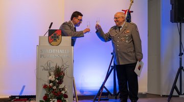 Zwei Männer in Uniform prosten sich auf einer kleinen Bühne mit Sektgläsern zu.