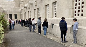 Jugendliche stehen in einer unregelmäßigen Reihe vor einem großen Gebäude und schauen durch Fenster am Boden hinein und unterhalten sich