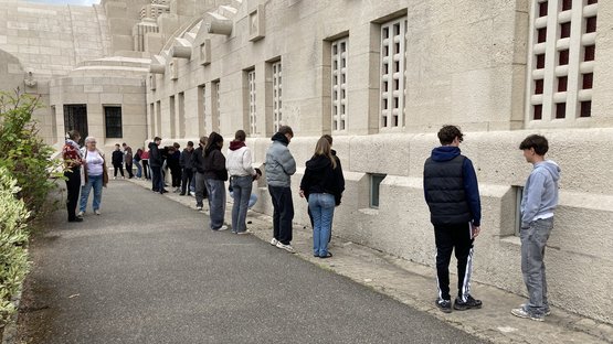Jugendliche stehen in einer unregelmäßigen Reihe vor einem großen Gebäude und schauen durch Fenster am Boden hinein und unterhalten sich