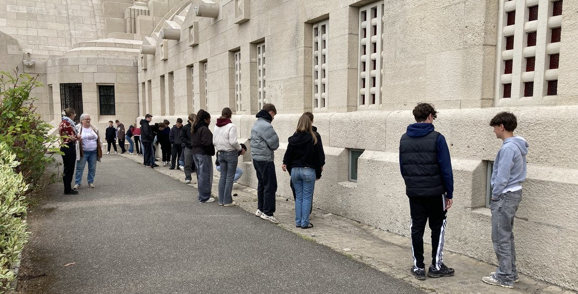 Jugendliche stehen in einer unregelmäßigen Reihe vor einem großen Gebäude und schauen durch Fenster am Boden hinein und unterhalten sich