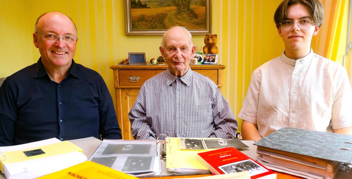 Drei Männer unterschiedlichen Alters nebeneinander am Tisch, vor ihnen liegen Bücher und Dokumente