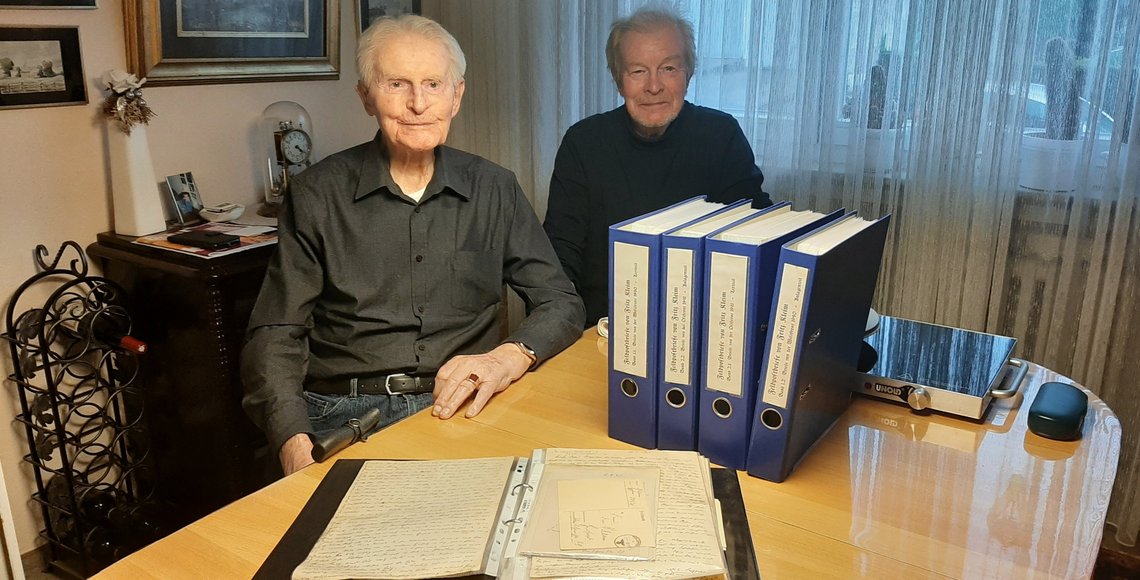 Zwei Männer nebeneinander am Tisch; auf dem Tisch stehen mehrere große Ordner.