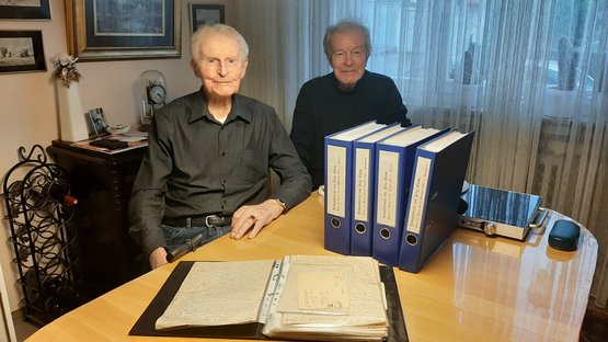 Zwei Männer nebeneinander am Tisch; auf dem Tisch stehen mehrere große Ordner.